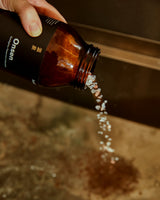 A hand holds an amber brown bottle labeled Onsen, pouring white granules into a sink over a brown surface.