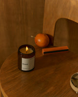 A lit candle in a dark amber glass jar sits on a wooden table, with a wooden chair back and an orange in the background. The label reads 'Strand'.
