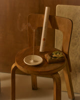 A wooden chair in a warm-toned interior setting, styled with a white incense holder, and smoking strand incense stick.