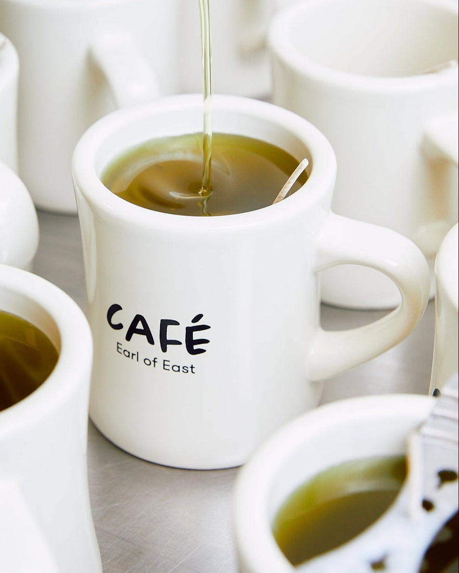 Iced Matcha Scented Candle in Mug, 300ml by Earl of East, lit, on a café table with coffee cups