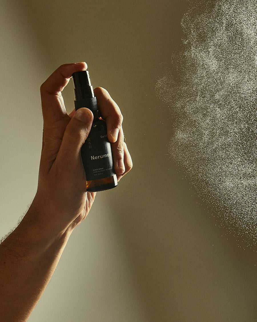 A hand holding a black spray bottle and spraying a fine mist toward the right against a warm beige background.