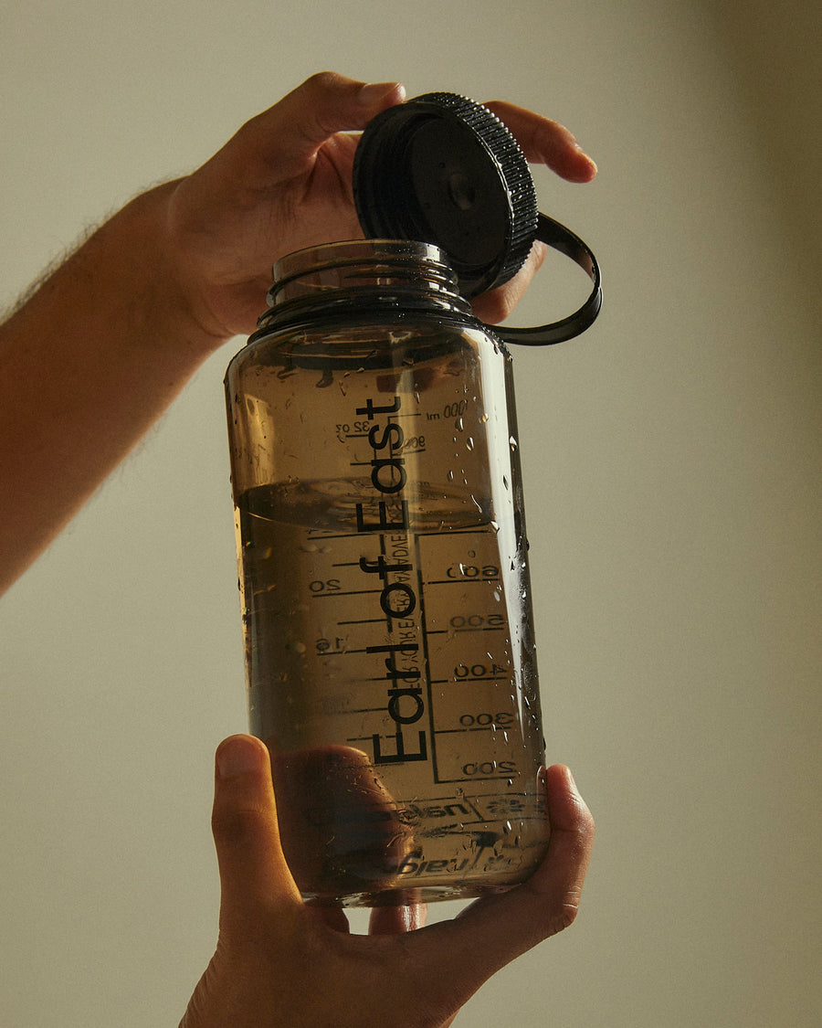 A person holds a clear plastic water bottle with a black cap and handle, showing the brand name 'Earth of East' printed vertically on the side. The bottle is filled with water and photographed against a plain beige background.
